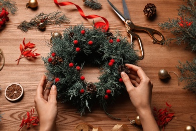 Florist making beautiful Christmas wreath with berries and pine cones at wooden table, above view