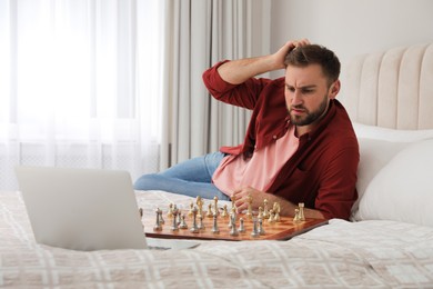 Thoughtful young man playing chess with partner through online video chat on bed at home