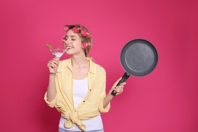 Young housewife with frying pan and glass of martini on pink background