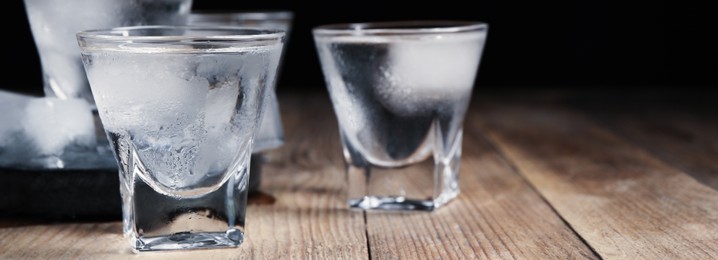 Image of Shot glasses of vodka with ice cubes on wooden table against black background, closeup view with space for text. Banner design
