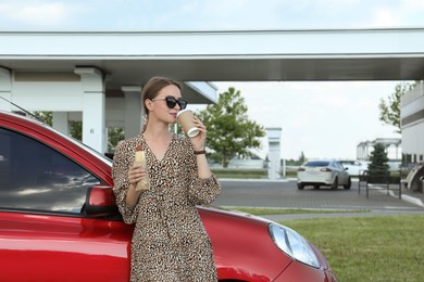 Beautiful young woman with hot dog drinking coffee near car at gas station