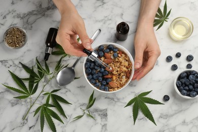 Top view of woman dripping THC tincture or CBD oil into oatmeal bowl at white marble table, closeup