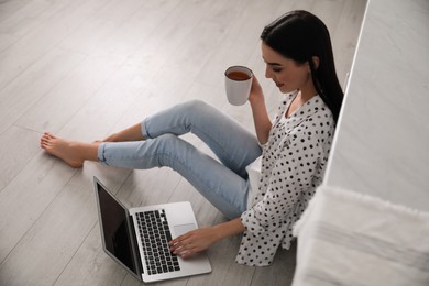 Happy woman with laptop and cup of drink sitting on warm floor indoors. Heating system
