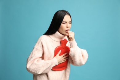 Ill woman with hot water bottle coughing on light blue background