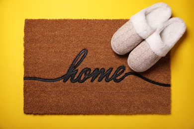 Photo of Doormat with word Home and slippers on yellow background, flat lay