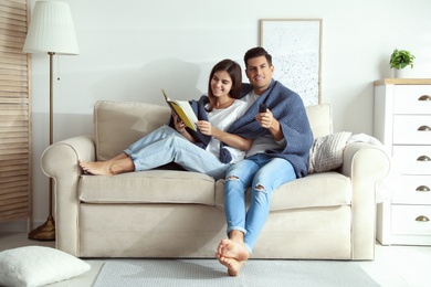 Happy couple covered with warm dark blue plaid reading book on sofa at home
