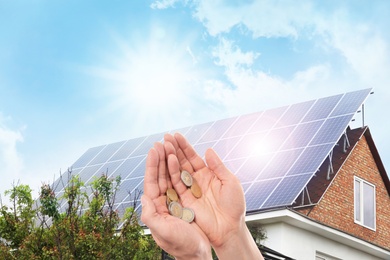 Man holding coins against house with installed solar panels. Renewable energy and money saving