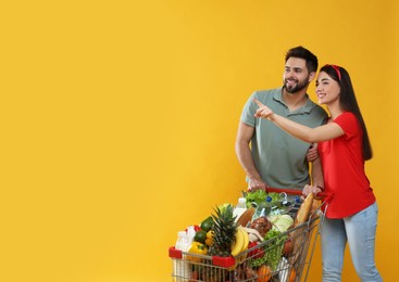 Young couple with shopping cart full of groceries on yellow background