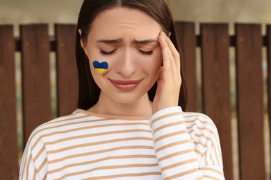 Photo of Sad young woman with drawing of Ukrainian flag on face outdoors