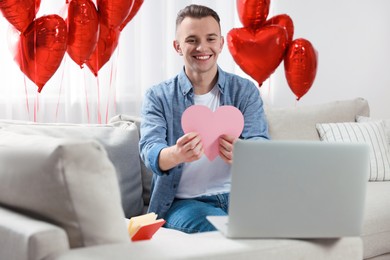 Photo of Valentine's day celebration in long distance relationship. Man holding pink paper heart while having video chat with his girlfriend via laptop