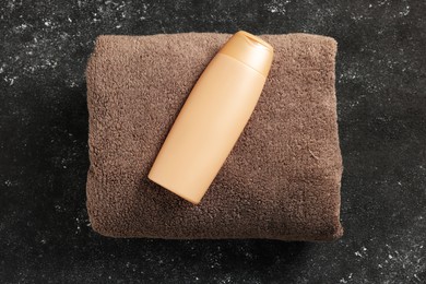 Soft brown terry towel and cosmetic bottle on black textured background, top view