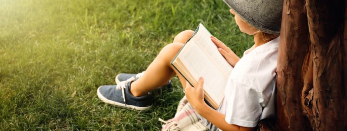 Cute little boy reading book on green grass near tree in park, space for text. Banner design