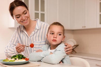 Mother feeding her daughter in kitchen. Little girl refusing to eat dinner