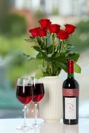 Bottle, glasses of red wine and vase with roses on white table. Romantic date