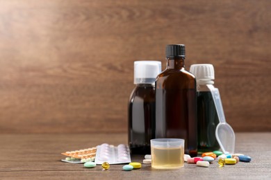 Photo of Bottles of syrup, measuring cup, dosing spoon and pills on wooden table, space for text. Cold medicine