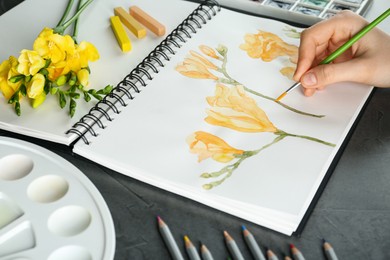 Woman drawing beautiful freesia flowers in sketchbook at black table, closeup