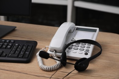 Stationary phone and headset near modern computer on wooden desk indoors. Hotline service