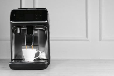 Modern espresso machine pouring coffee into cup on white wooden table near light wall. Space for text