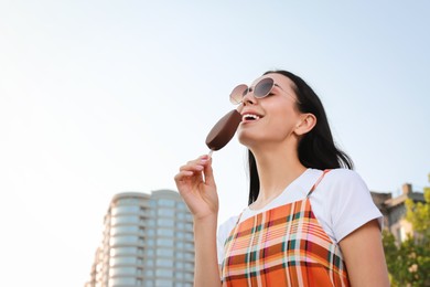 Beautiful young woman eating ice cream glazed in chocolate on city street, low angle view. Space for text