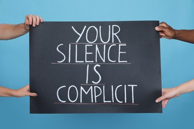Photo of People holding sign with phrase Your Silence Is Complicit on light blue background, closeup. Racism concept