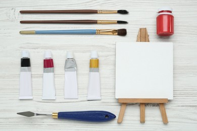 Easel with blank canvas, brushes and paints on white wooden table, flat lay. Space for text