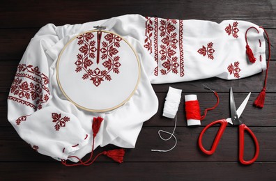 Shirt with red embroidery design in hoop, needle, scissors and threads on wooden table, flat lay. National Ukrainian clothes