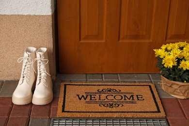 Door mat with word Welcome, stylish boots and beautiful flowers near entrance