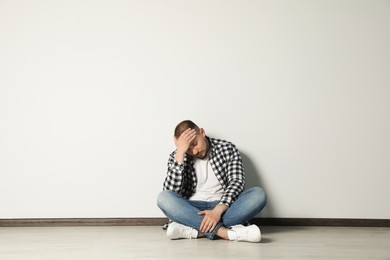 Photo of Sad young man sitting on floor near white wall indoors, space for text