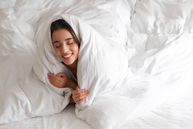 Cheerful young woman covered with warm white blanket lying in bed. Space for text