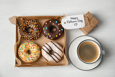Tag with phrase HAPPY FATHER'S DAY, donuts, coffee and gift on white wooden table, flat lay