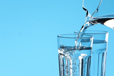 Photo of Pouring water from jug into glass on light blue background, closeup. Space for text