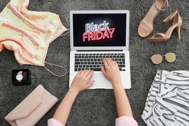 Woman surrounded by clothes and accessories using laptop with Black Friday announcement on grey carpet, top view