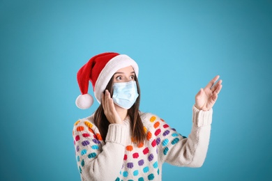 Surprised woman in Santa hat and medical mask pointing on light blue background