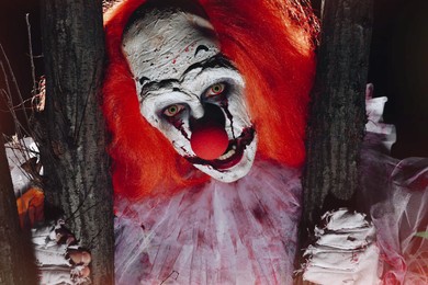 Terrifying clown hiding behind trees outdoors at night, closeup. Halloween party costume