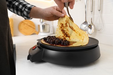 Photo of Man cooking delicious crepe with jam on electric pancake maker in kitchen, closeup