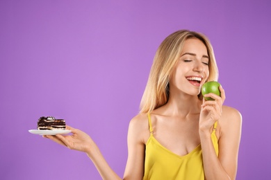 Woman choosing between cake and healthy apple on violet background