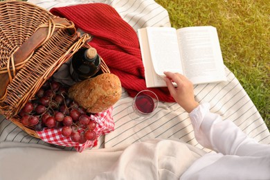 Woman with glass of wine, book and picnic basket on green grass, above view