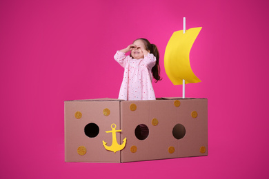 Little child playing with ship made of cardboard box on pink background