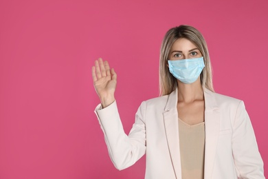 Woman in protective mask showing hello gesture on pink background, space for text. Keeping social distance during coronavirus pandemic