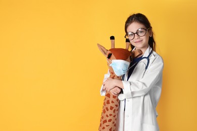 Little girl with eyeglasses and stethoscope as doctor hugging toy giraffe on yellow background, space for text. Pediatrician practice