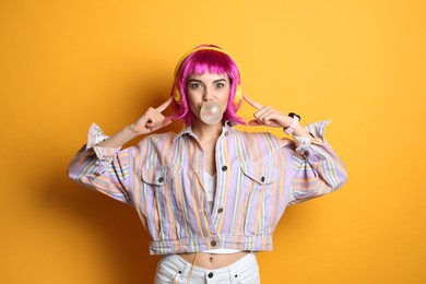 Fashionable young woman in colorful wig with headphones blowing bubblegum on yellow background