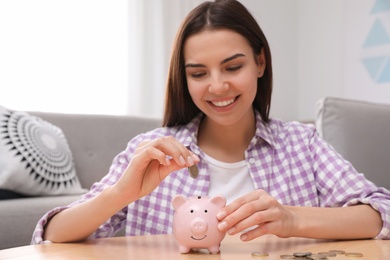 Happy young woman putting coin into piggy bank at home