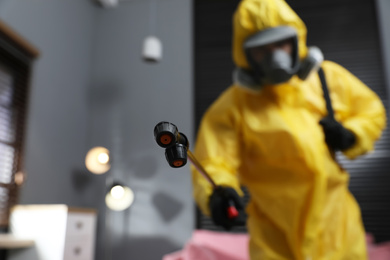 Pest control worker in protective suit indoors, focus on insecticide sprayer