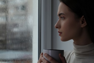 Melancholic young woman with cup of drink near window on rainy day, space for text. Loneliness concept