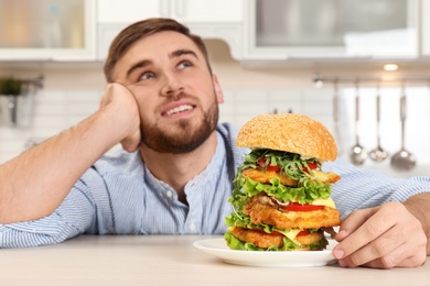 Photo of Young dreamy man holding plate with huge burger on table