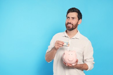Happy man putting money into piggy bank on light blue background, space for text