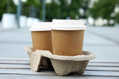 Takeaway paper coffee cups with plastic lids in cardboard holder on wooden bench outdoors