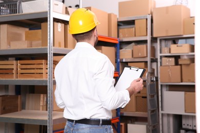 Young man with clipboard near rack of cardboard boxes at warehouse, back view