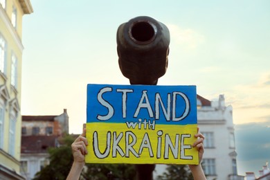 Woman holding poster in colors of national flag and words Stand With Ukraine near broken military tank outdoors, closeup
