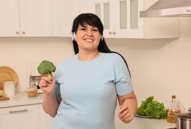 Happy overweight woman with headphones and broccoli in kitchen. Healthy diet